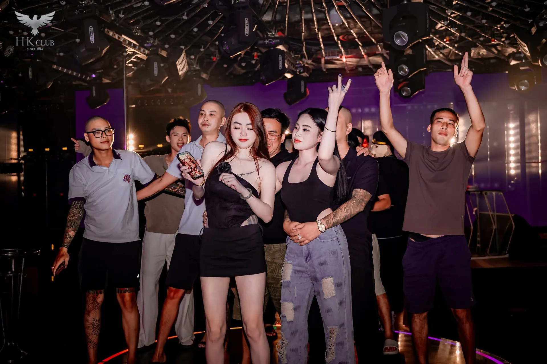 Nhóm bạn giơ tay trên sân khấu tròn nền tím tại HK Club